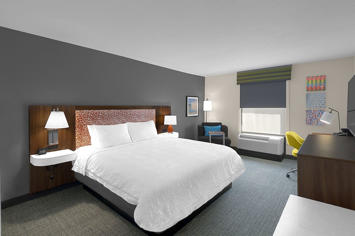 A hotel room with a large bed, bedside tables with lamps, a desk with a chair, an air conditioning unit, and a window with blinds.