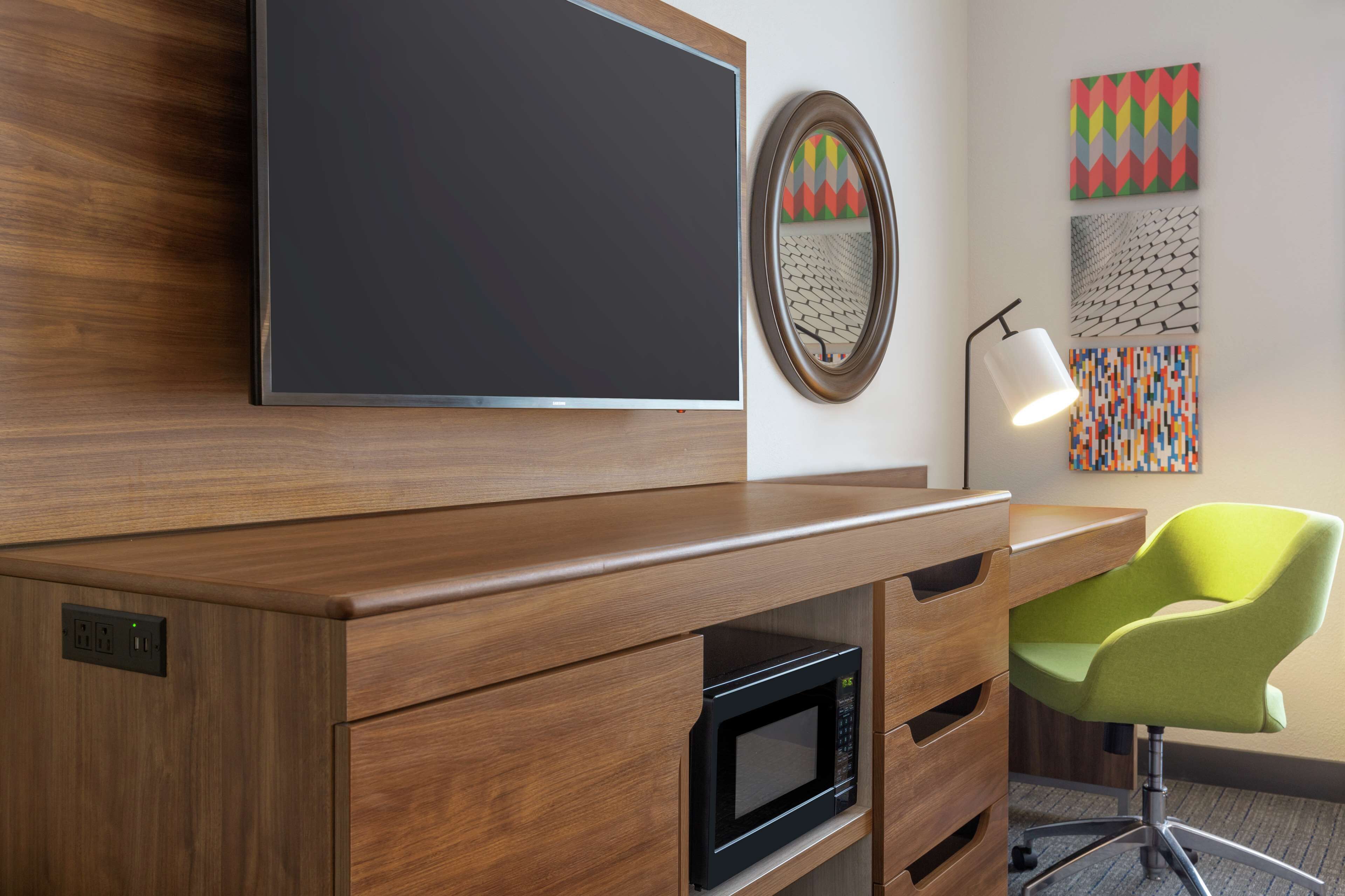 A modern office space with a wooden desk, a built-in microwave, a green chair, a flat-screen TV, and decorative wall art.