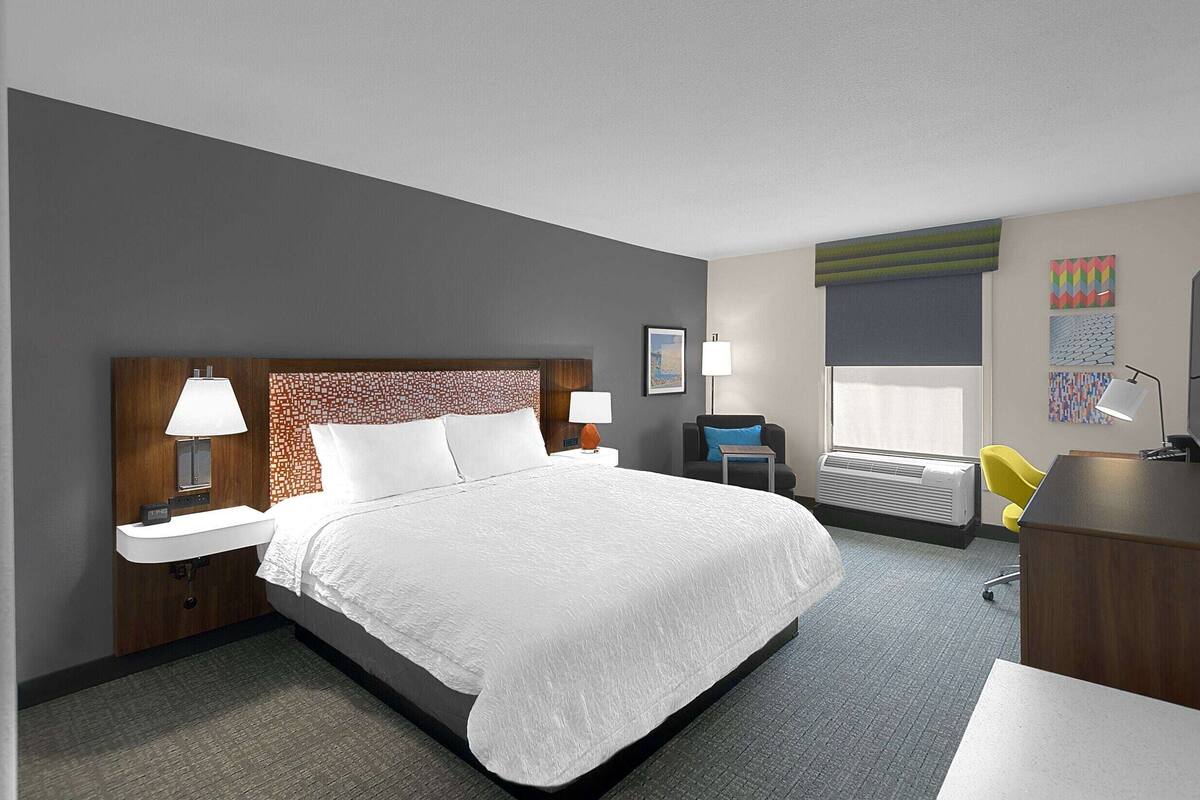 A hotel room with a large bed, bedside lamps, a desk, a chair, and a window with blinds.
