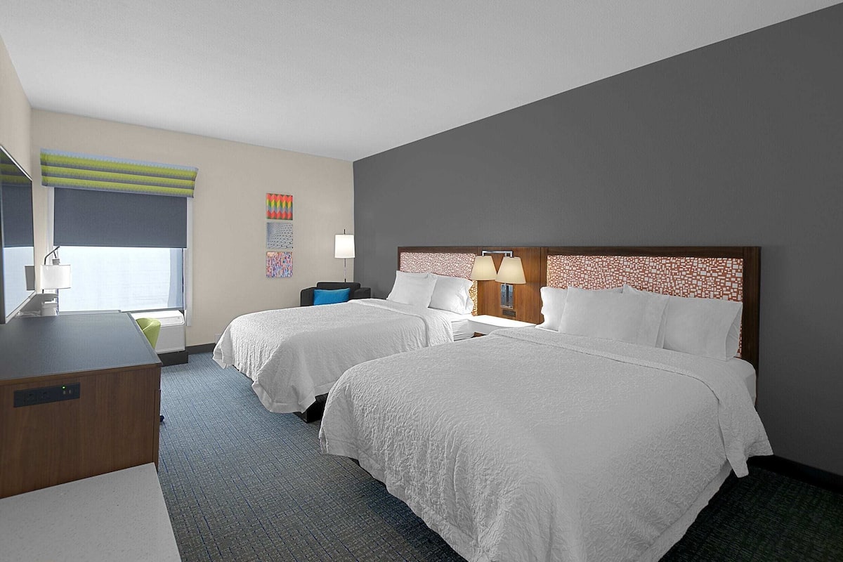 A hotel room with two beds, a desk, a chair, a lamp, and a window with a view.