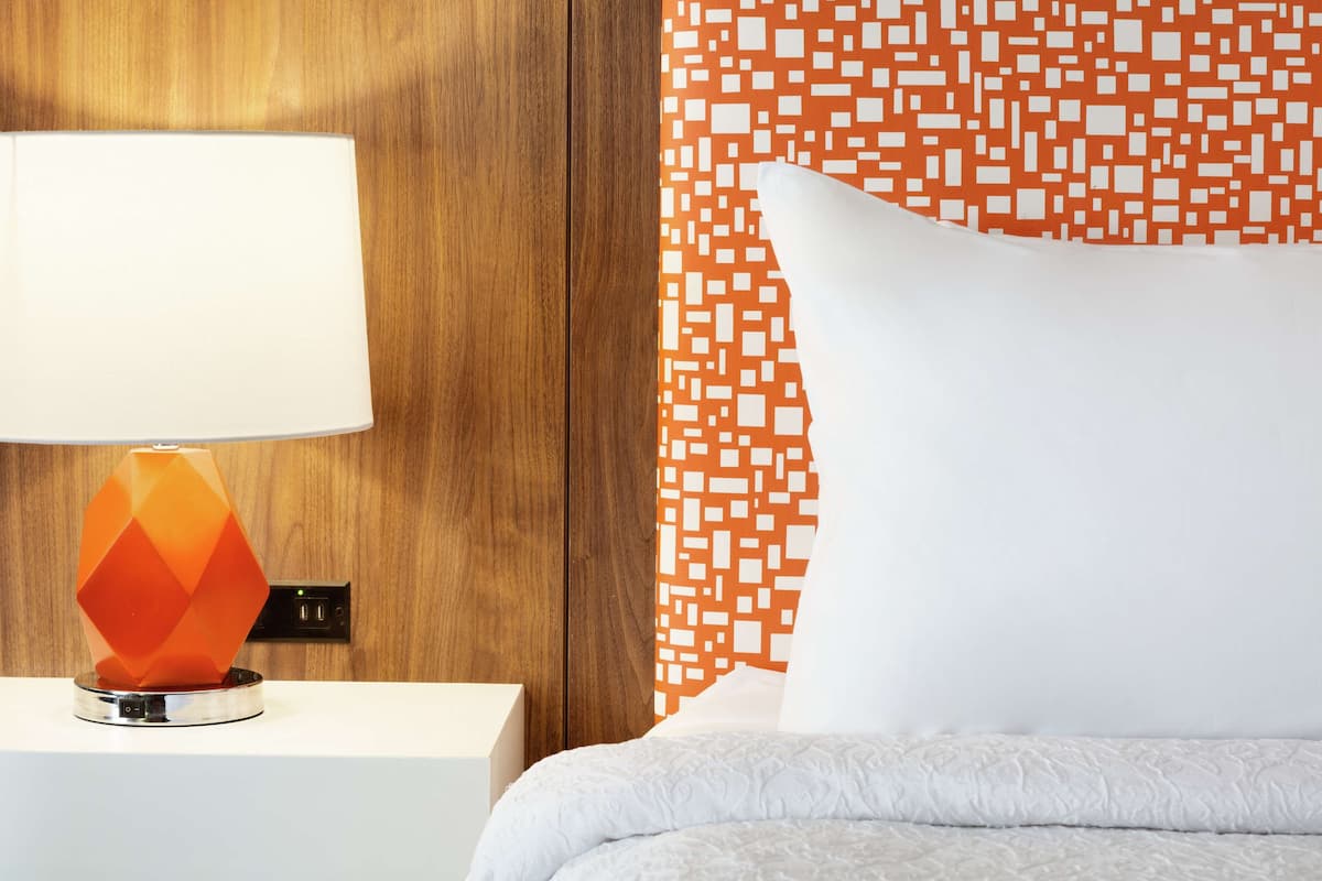 A neatly made bed with a white pillow and a white duvet, a modern orange and white patterned wall, and a white nightstand with a lamp.