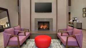 A modern living room with a fireplace, two pink armchairs, a red ottoman, and a flat-screen TV mounted above the fireplace. - Hampton Inn & Suites Houston-Cypress Station (Houston)