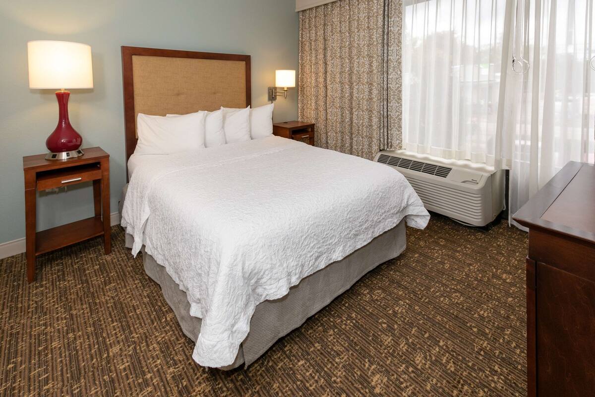 Een hotelkamer met een groot bed, een bedlampje, een airconditioning en een raam met gordijnen.