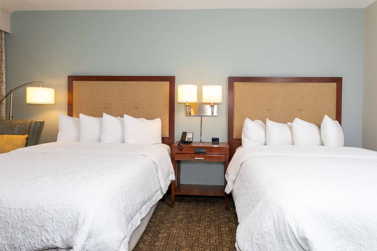 Een hotelkamer met twee bedden, elk voorzien van witte lakens en een houten hoofdbord. Tussen de bedden staat een nachtkastje met een lamp en een telefoon.