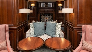 Ein Zimmer mit einer holzverkleideten Wand, einer Couch mit gemusterten Kissen, zwei runden Holztischen und einem Kamin mit aufgehängten Bildern darüber.