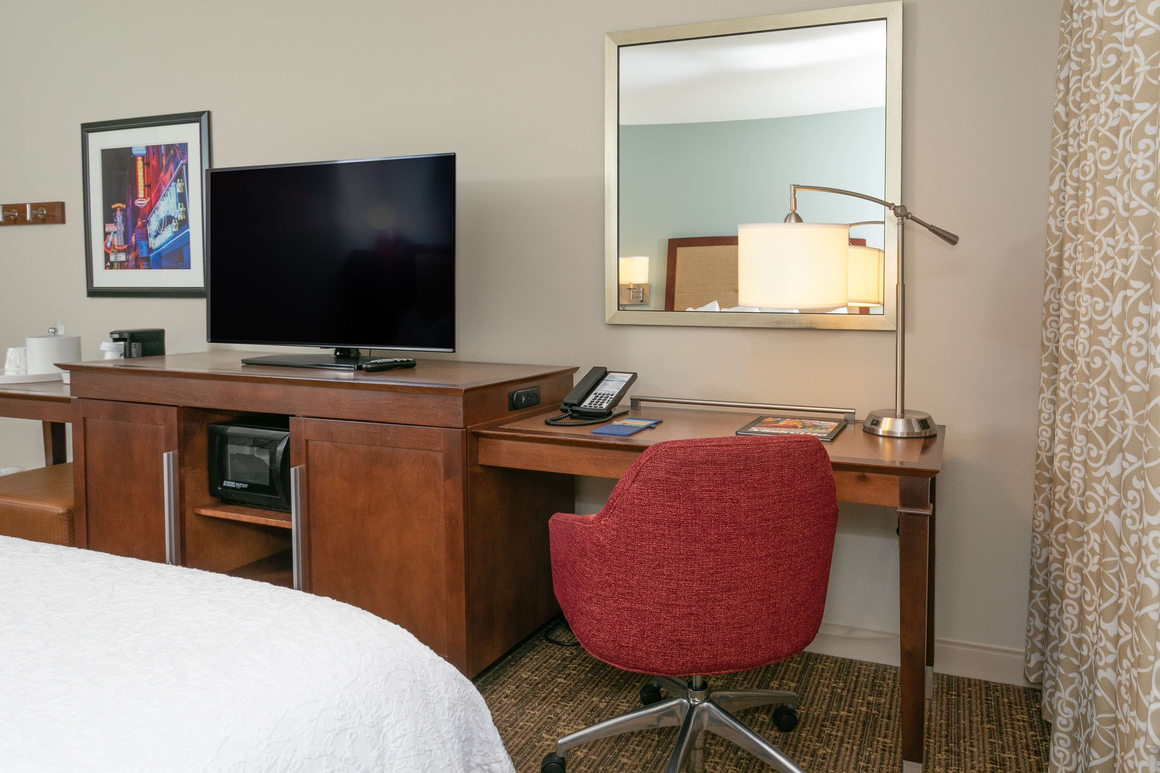 Ein Hotelzimmer mit einem Bett, einem Schreibtisch mit Fernseher, einem roten Sessel, einem Spiegel und einer Lampe.