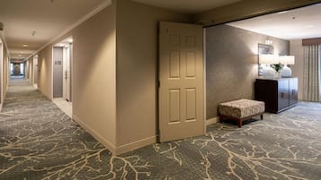 A hotel corridor with carpeted floors, a closed door, a bench, and a cabinet with a lamp.