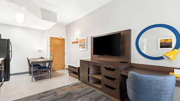 A modern living room with a flat-screen TV, a blue office chair, a wooden desk, and a dining area with a table and chairs.