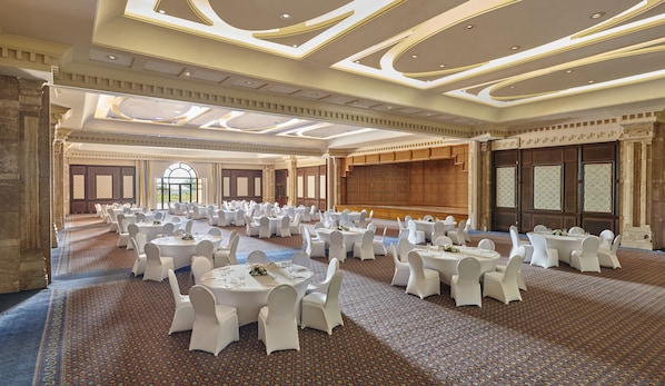 Un amplio salón de banquetes con mesas redondas y sillas blancas, alfombra estampada y un techo alto con iluminación empotrada.