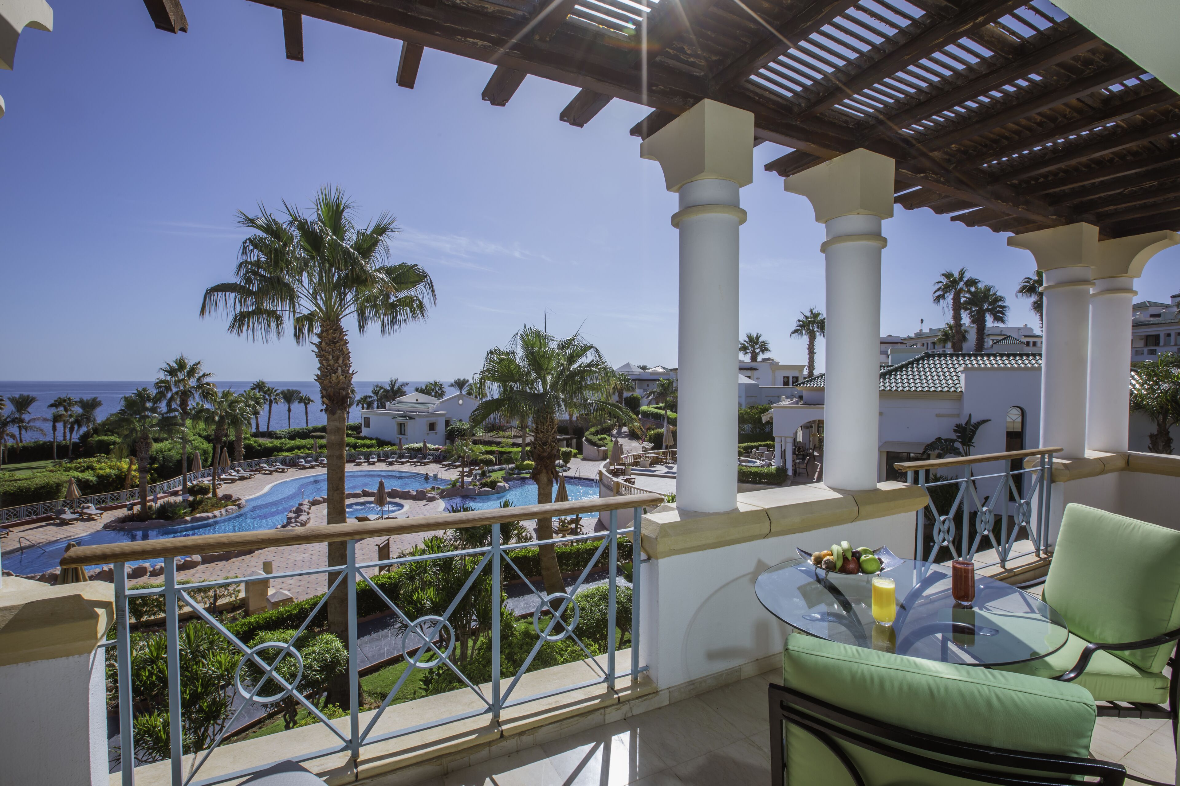 A balcony with a glass table, a green chair, and a view of a pool and palm trees.