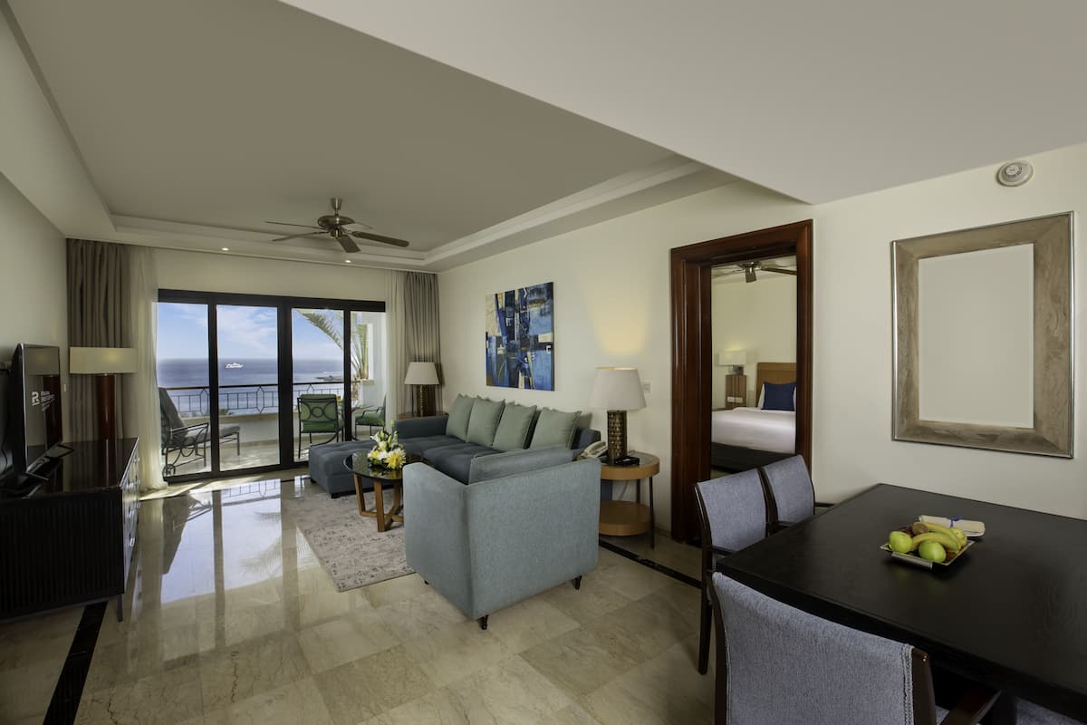 A spacious living room with a sofa, a dining table with fruit, a balcony with a view of the sea, and a ceiling fan.