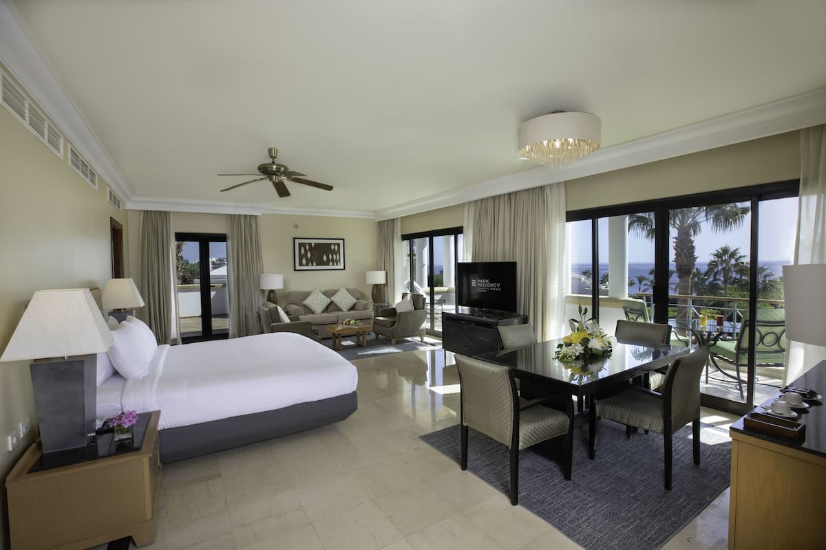 A hotel room with a large bed, a TV, a dining table with chairs, and a balcony with a view.