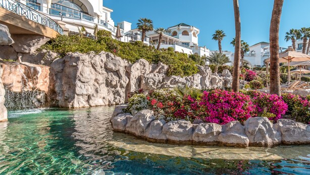 A clear pool with a rocky edge, surrounded by vibrant pink flowers and palm trees, with a white building in the background. - Park Regency Sharm El Sheikh Resort (Sharm El Sheikh)