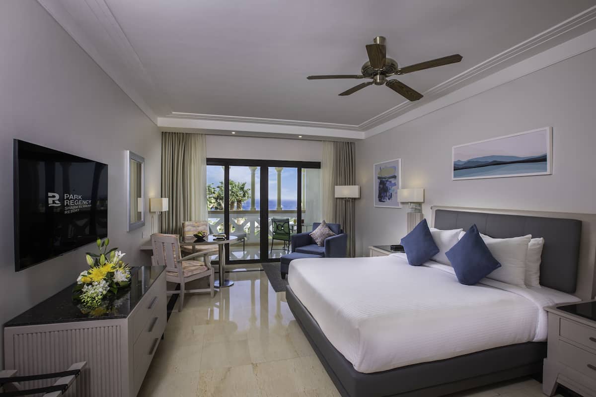 A hotel room with a large bed, a desk with a TV, a balcony with a view, and a ceiling fan.