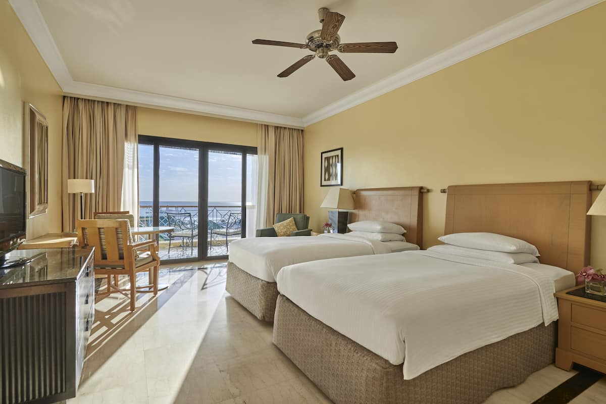 A hotel room with two beds, a TV, a desk, a chair, and a balcony with a view.