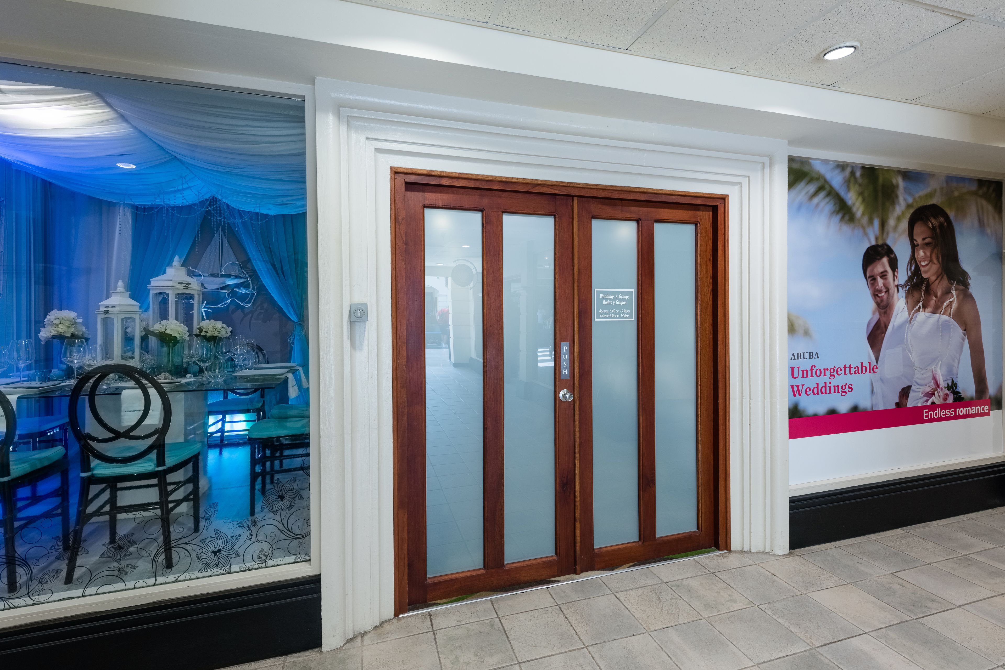 Entrada de hotel con puerta de madera, ventanal con vista a un comedor y un cartel anunciando bodas en Aruba.