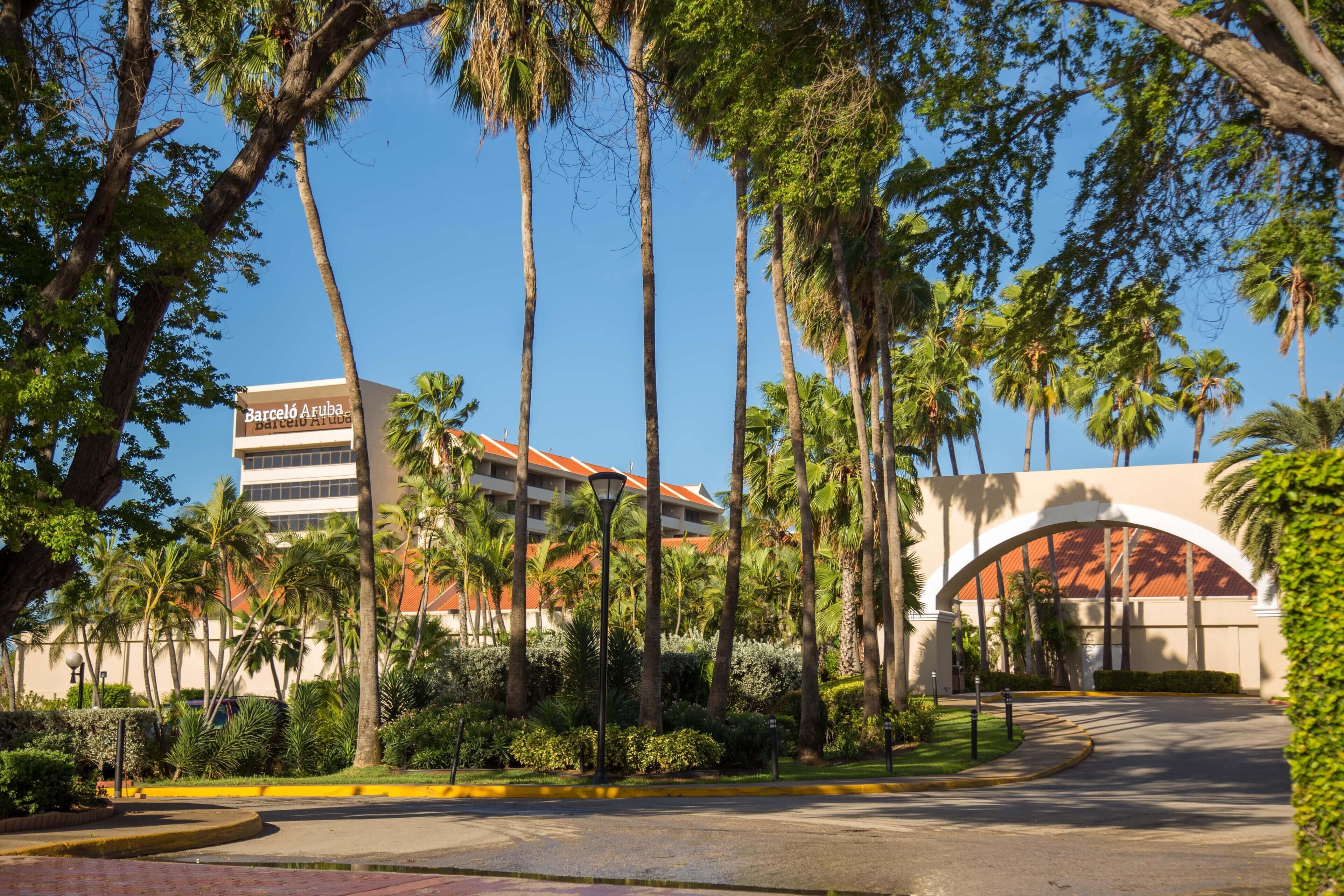 A hotel entrance with a palm tree-lined pathway, a building with a red roof, and a clear blue sky.