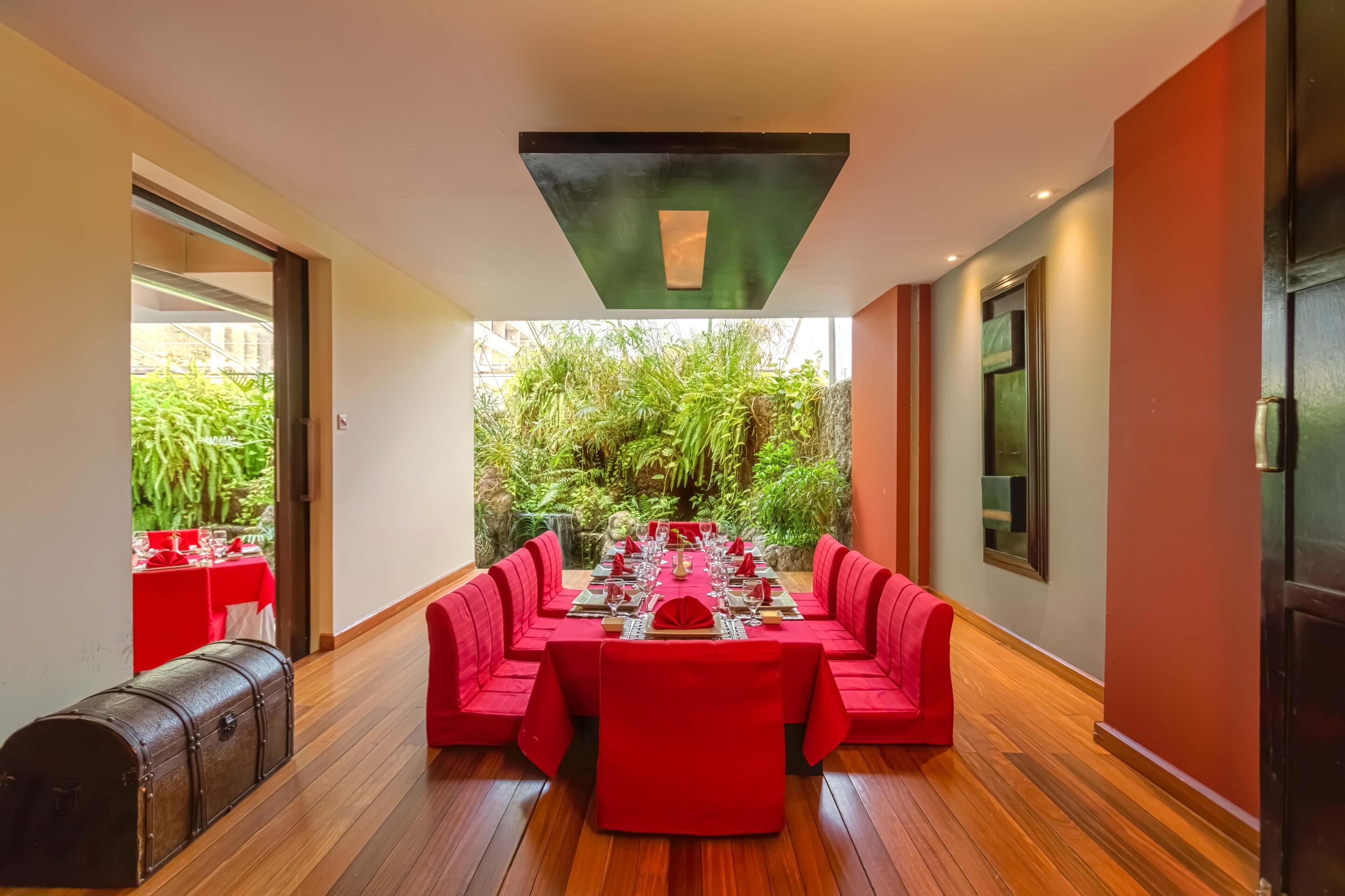 A dining area with a long table, red upholstered chairs, and a view of lush greenery outside.