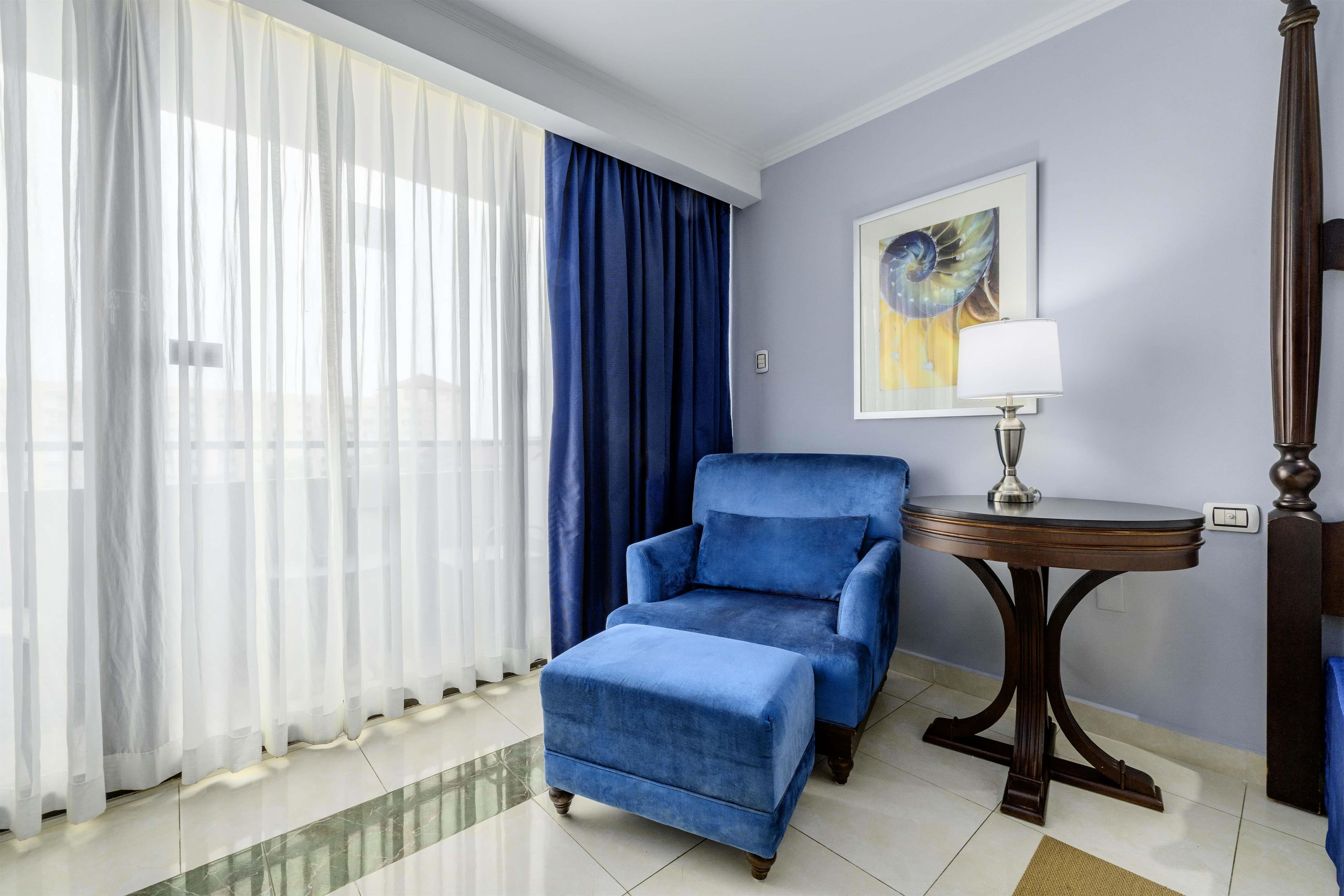 A room with a blue armchair, a small round table with a lamp, a painting on the wall, and sheer curtains.