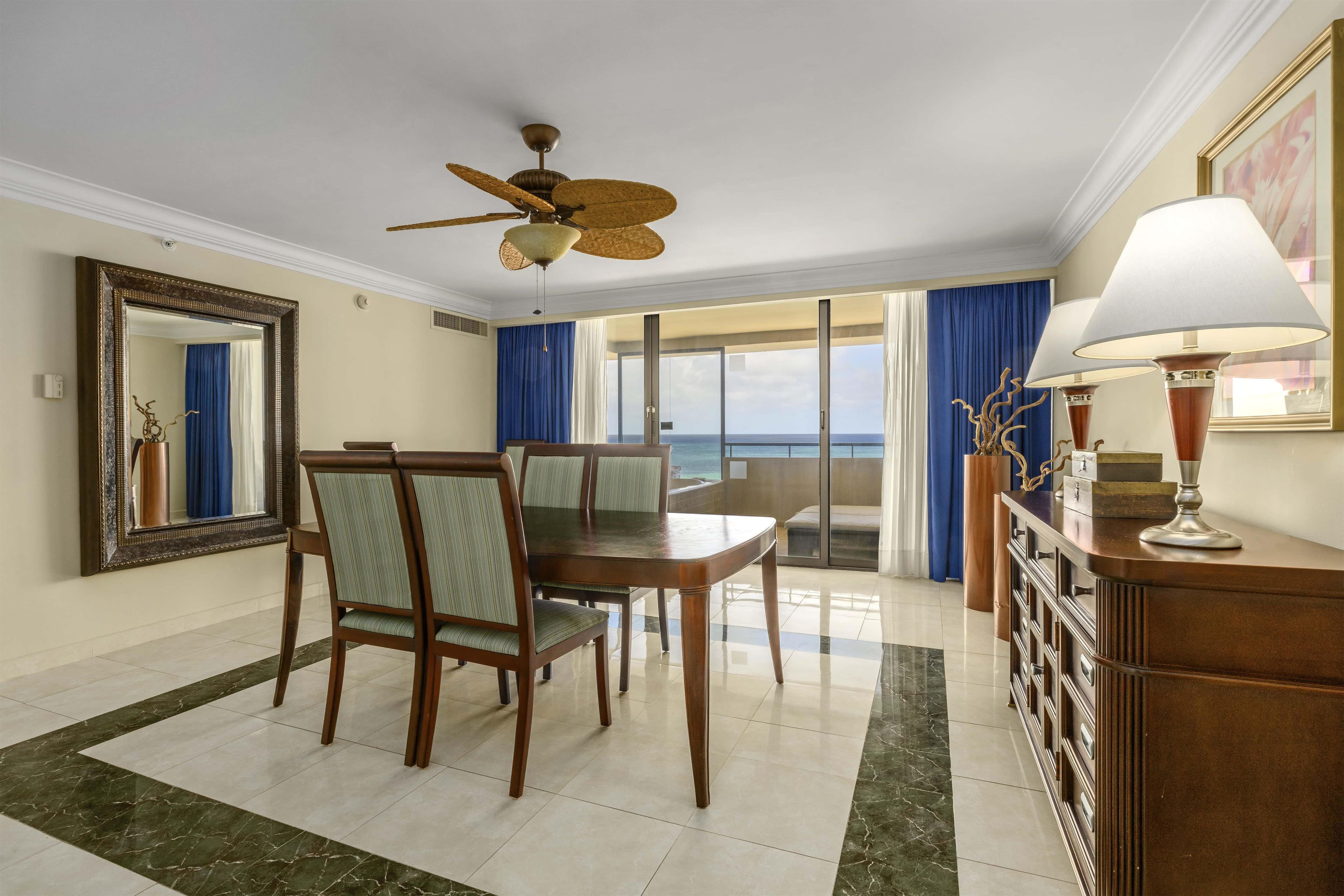 A hotel room with a dining area, a large mirror, a ceiling fan, a view of the ocean, and a lamp on a dresser.