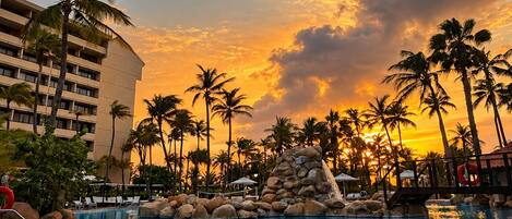 A resort pool area with a sunset view, palm trees, and a stone-tiled deck.
