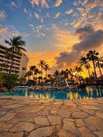 Zona de piscina en un resort con vista al atardecer, palmeras y un deck con baldosas de piedra.