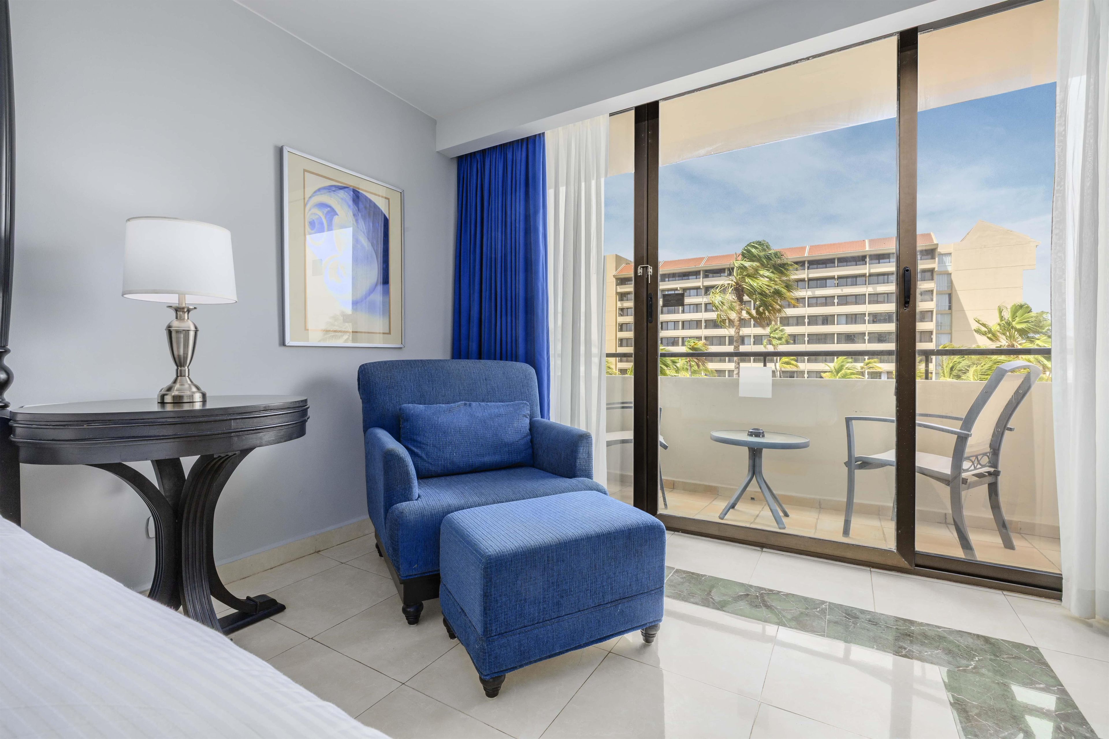 A hotel room with a bed, a desk, a chair, a small table, a balcony with a view of buildings and palm trees.
