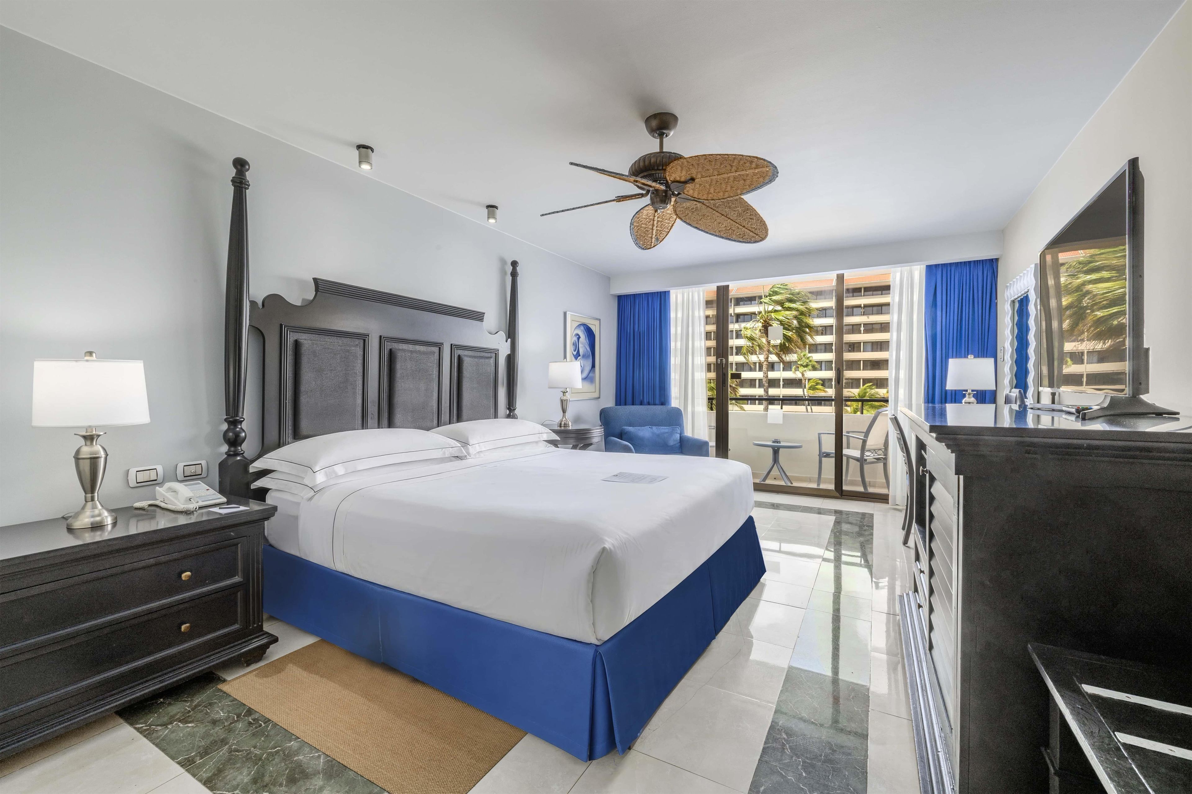 A hotel room with a bed, bedside table, lamp, and a view of the outside.