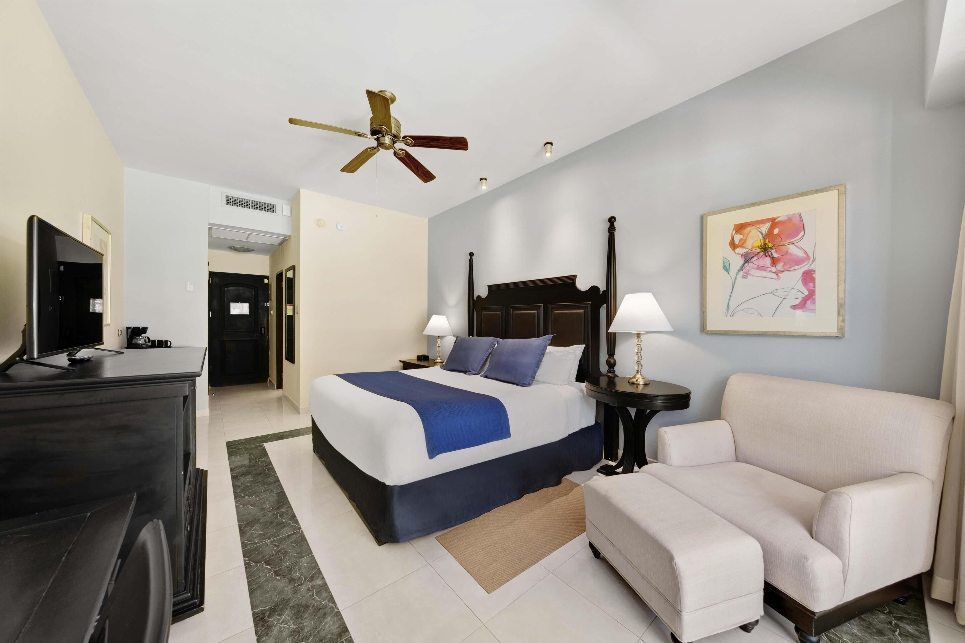 Habitación de hotel con cama, mesitas de noche, escritorio, silla, ventilador de techo y un cuadro en la pared.