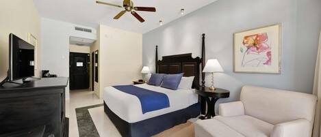 Habitación de hotel con cama, mesitas de noche, escritorio, silla, ventilador de techo y un cuadro en la pared.