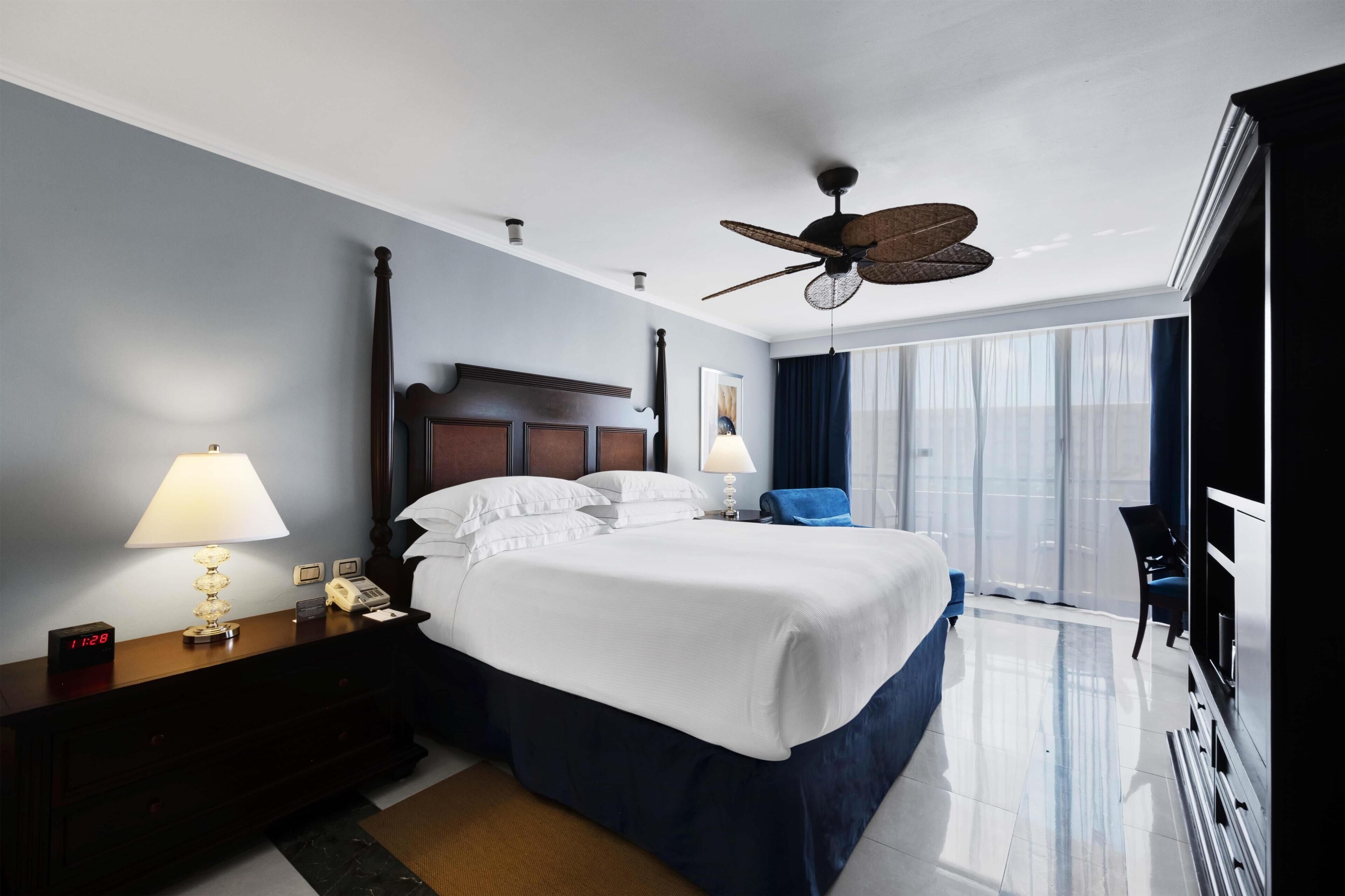 A hotel room with a large bed, two bedside tables with lamps, a ceiling fan, a TV, and a view of the city through the window.