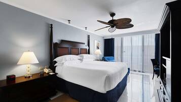 A hotel room with a large bed, two bedside tables with lamps, a ceiling fan, a TV, and a view of the city through the window.