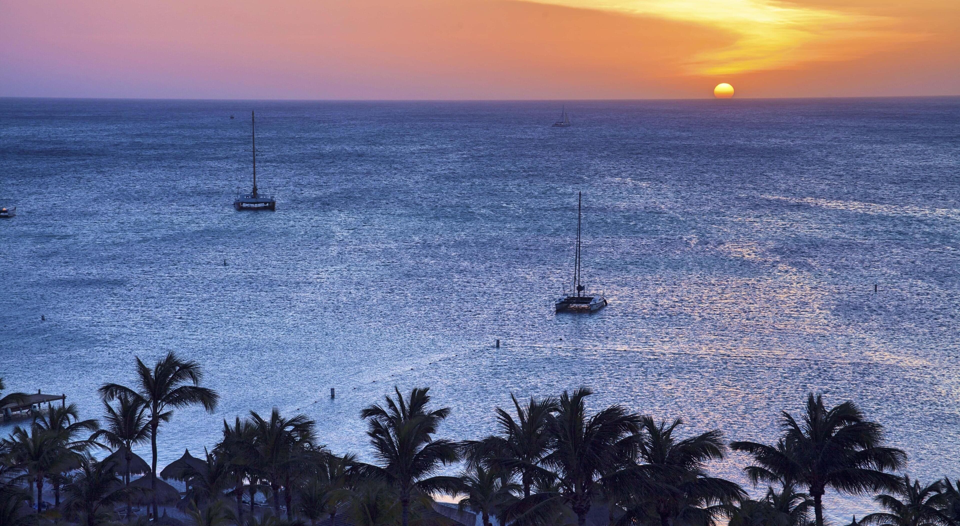 Atardecer sobre un mar tranquilo con veleros y palmeras.