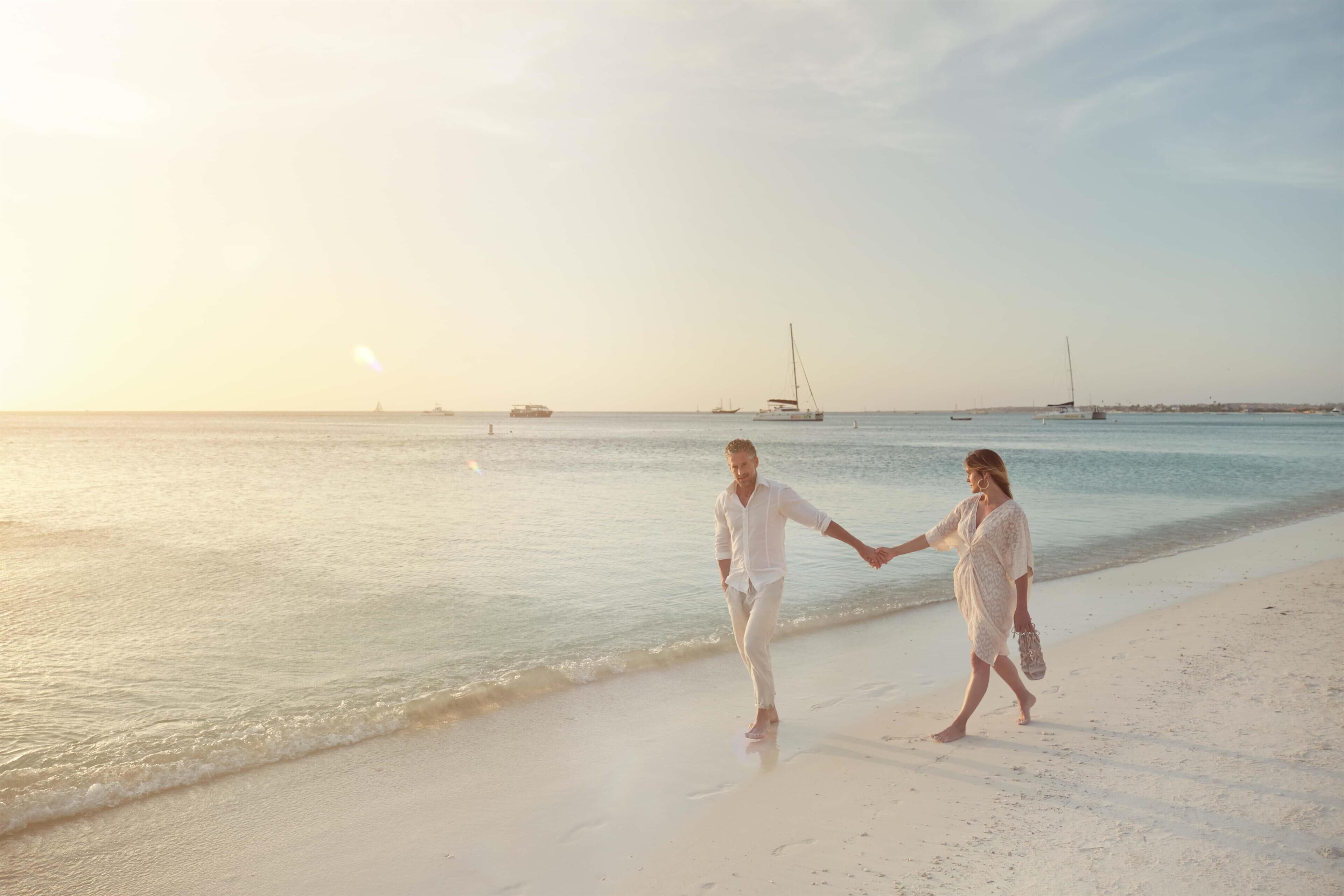 A couple walking on a beach with boats in the background.