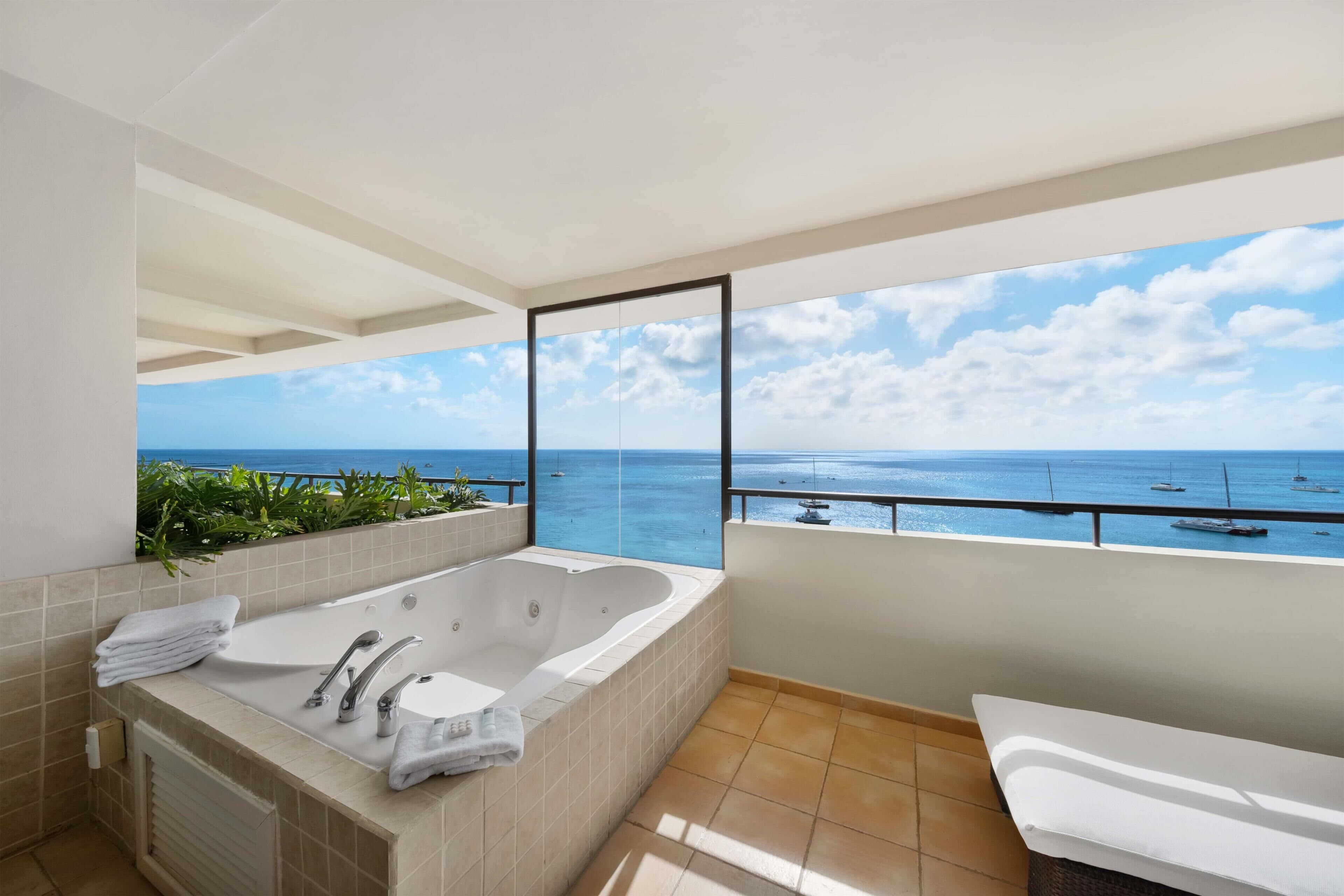 A bathroom with a large bathtub, a toilet, and a sink. There is a view of the ocean and boats through a large window.