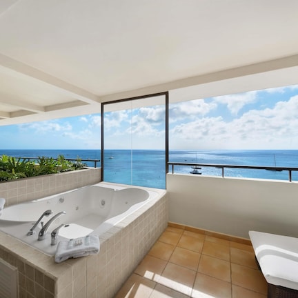 A bathroom with a large bathtub, a toilet, and a sink. There is a view of the ocean and boats through a large window.