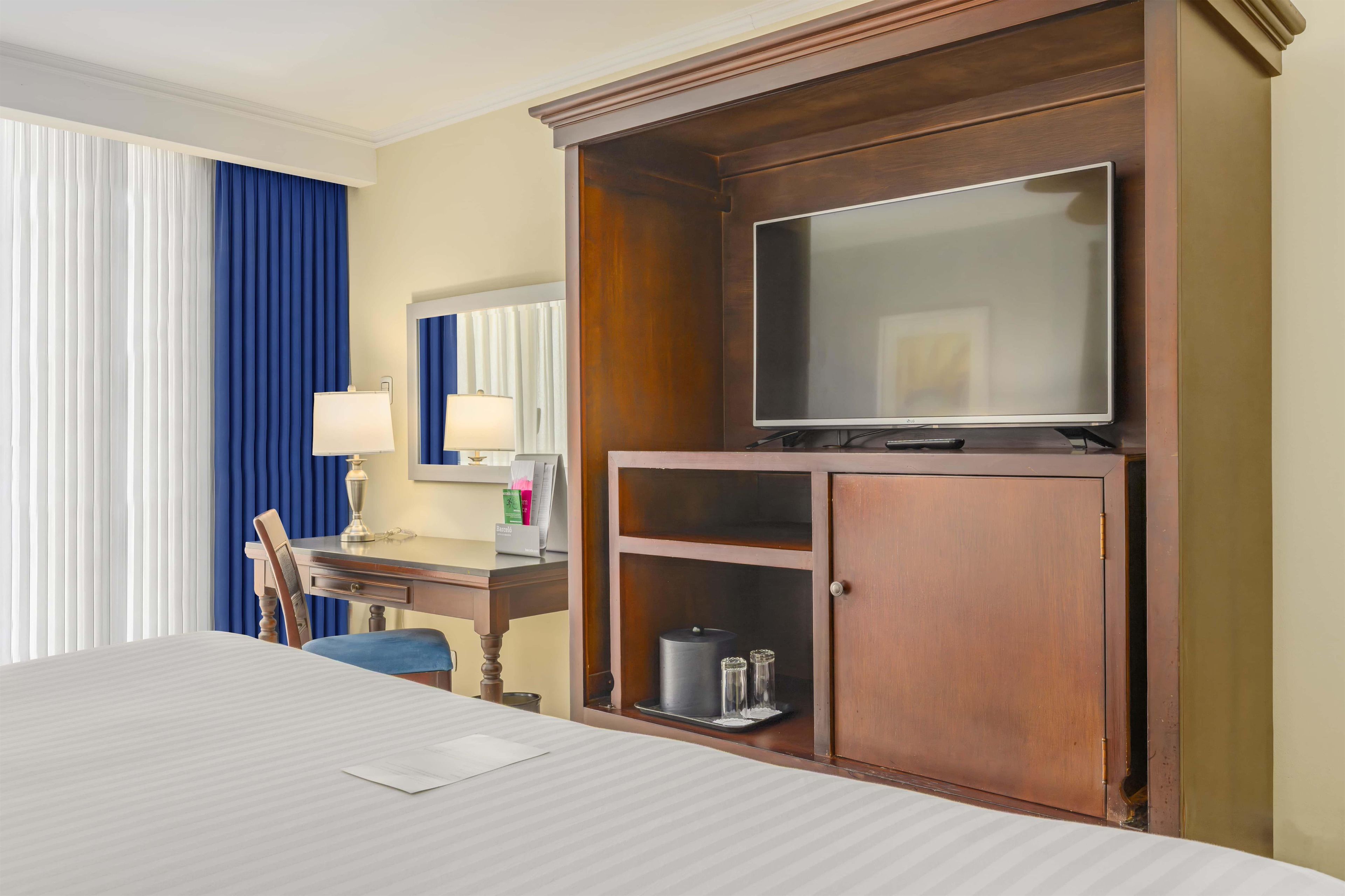 A hotel room with a bed, a desk with a lamp, a television on a wooden cabinet, and a window with curtains.