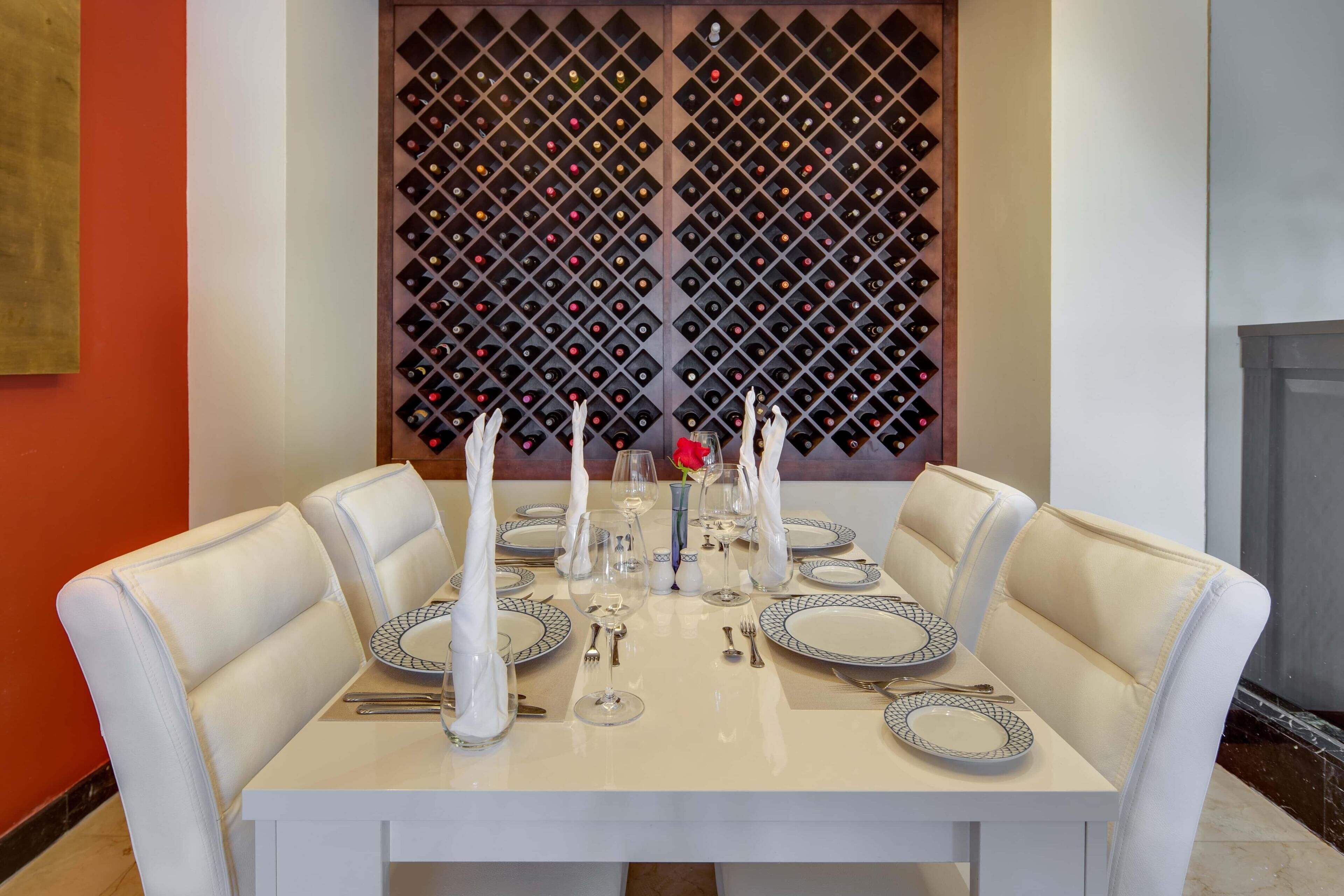 Una mesa de comedor dispuesta para seis personas, con sillas blancas, una bodega en el fondo y una pared roja.