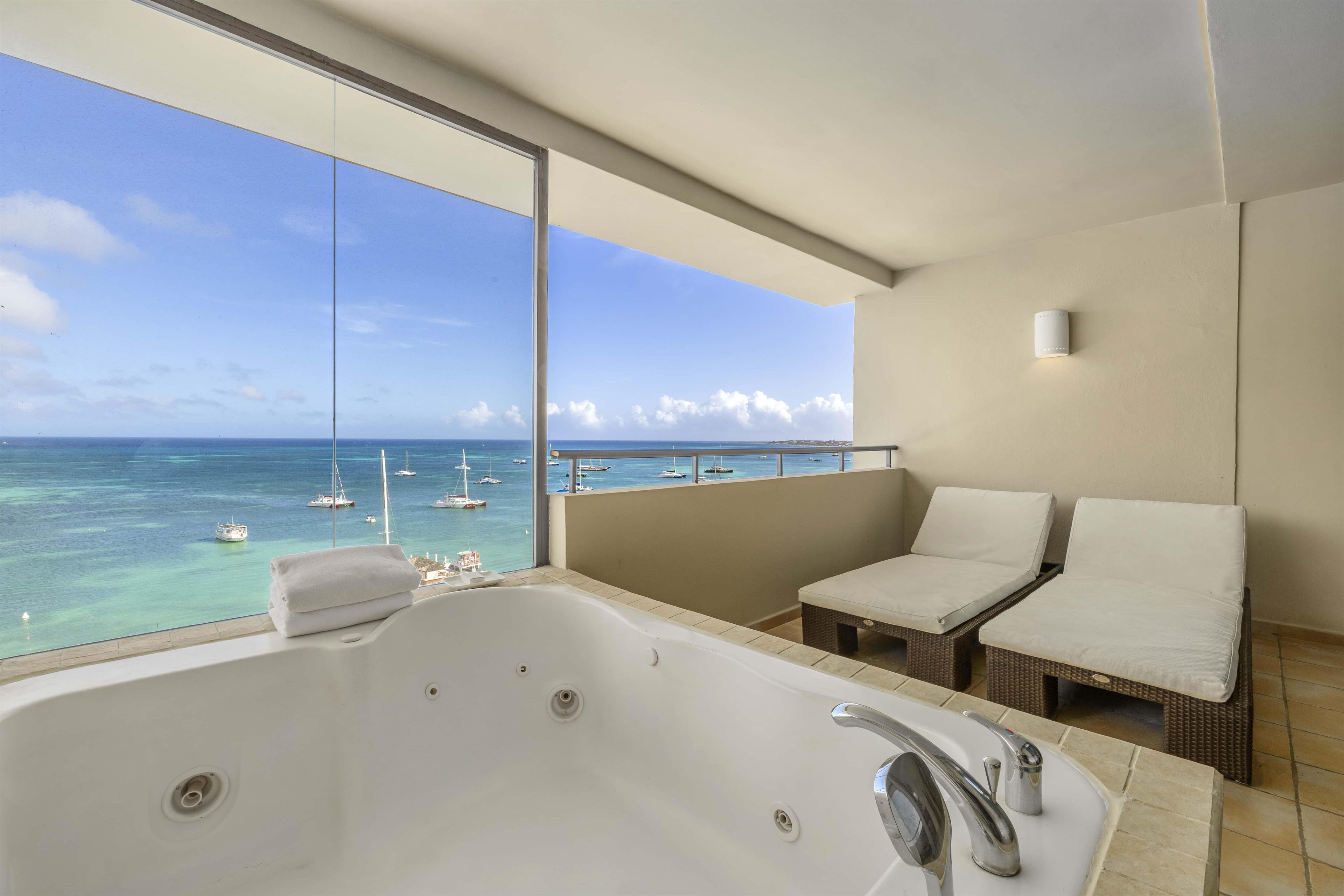 Un balcon avec vue sur la mer et les bateaux, une baignoire blanche et deux fauteuils blancs avec des coussins.