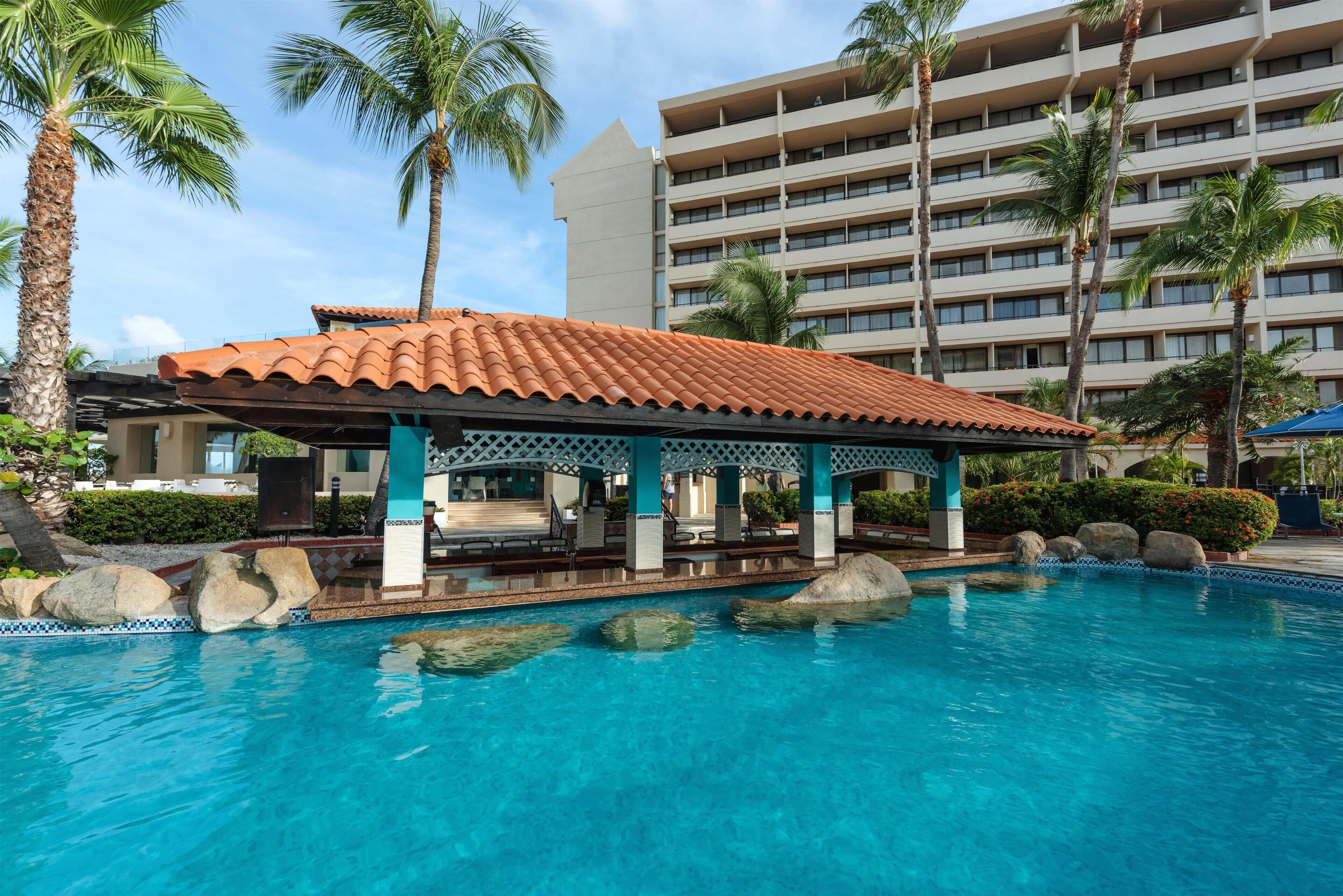 A hotel with a pool, palm trees, and a covered seating area.