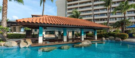 A hotel with a pool, palm trees, and a covered seating area.