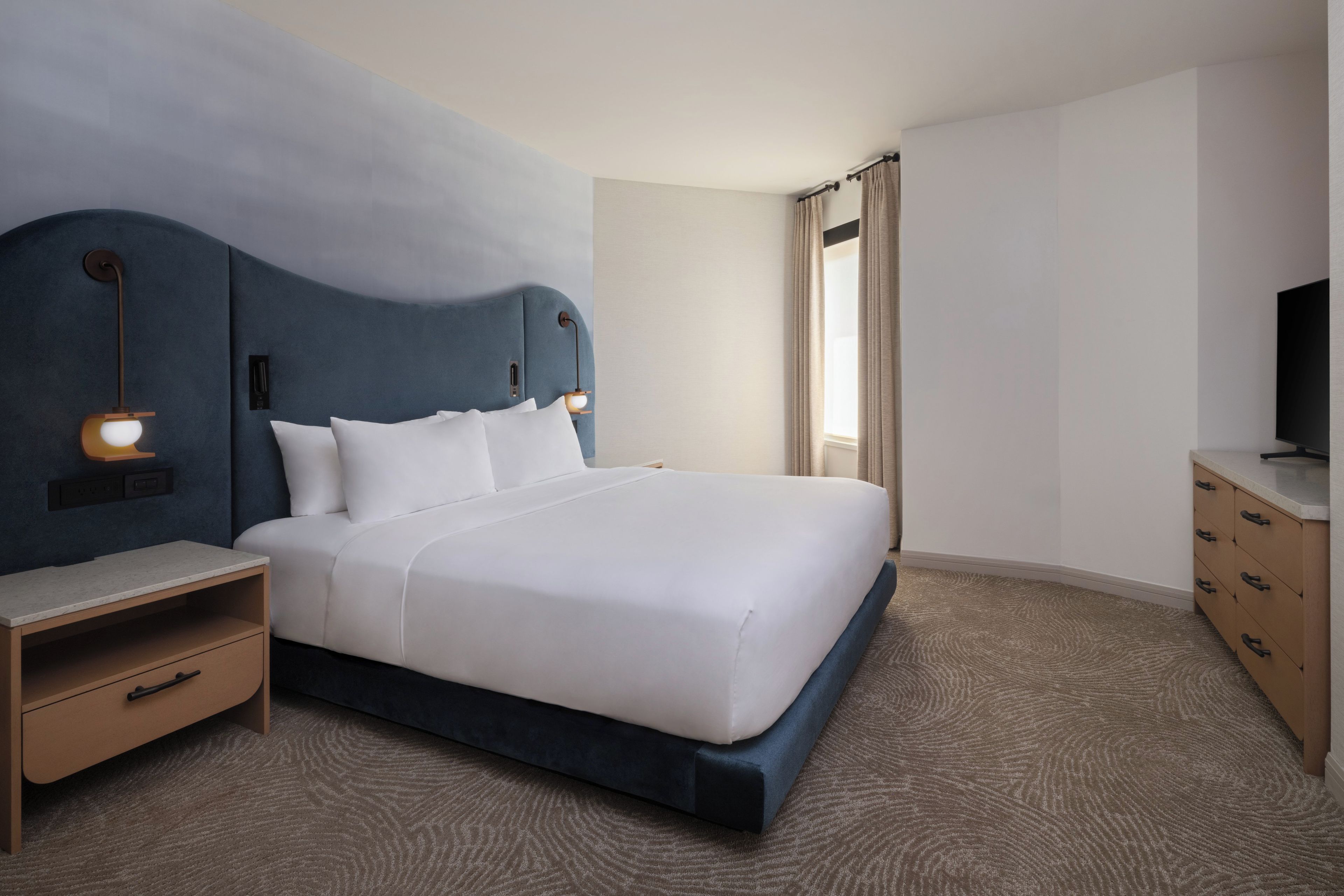Una habitación de hotel con una cama grande, mesitas de noche, un televisor y una ventana con cortinas.