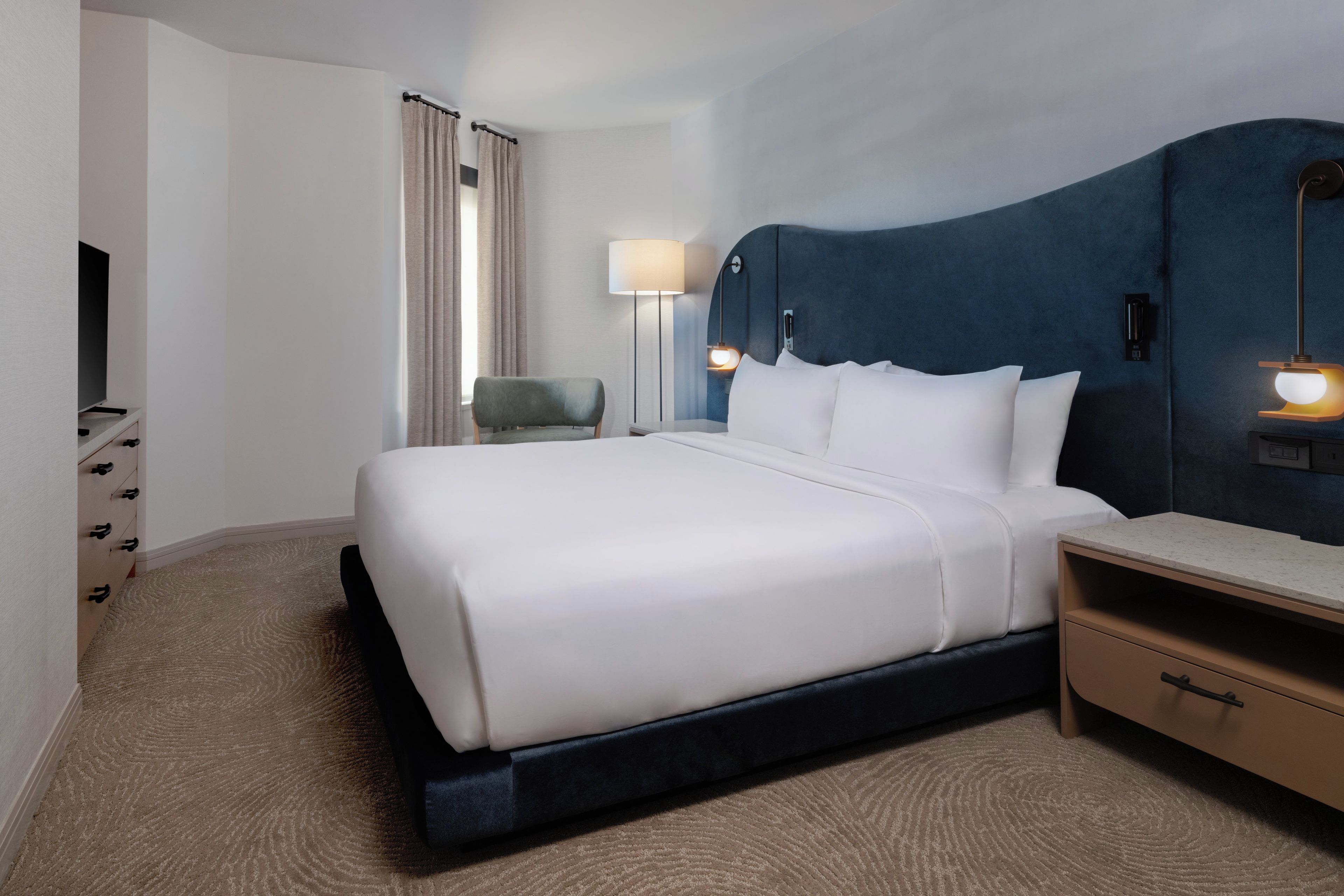 A hotel room with a bed, a chair, a desk, a lamp, and a television.