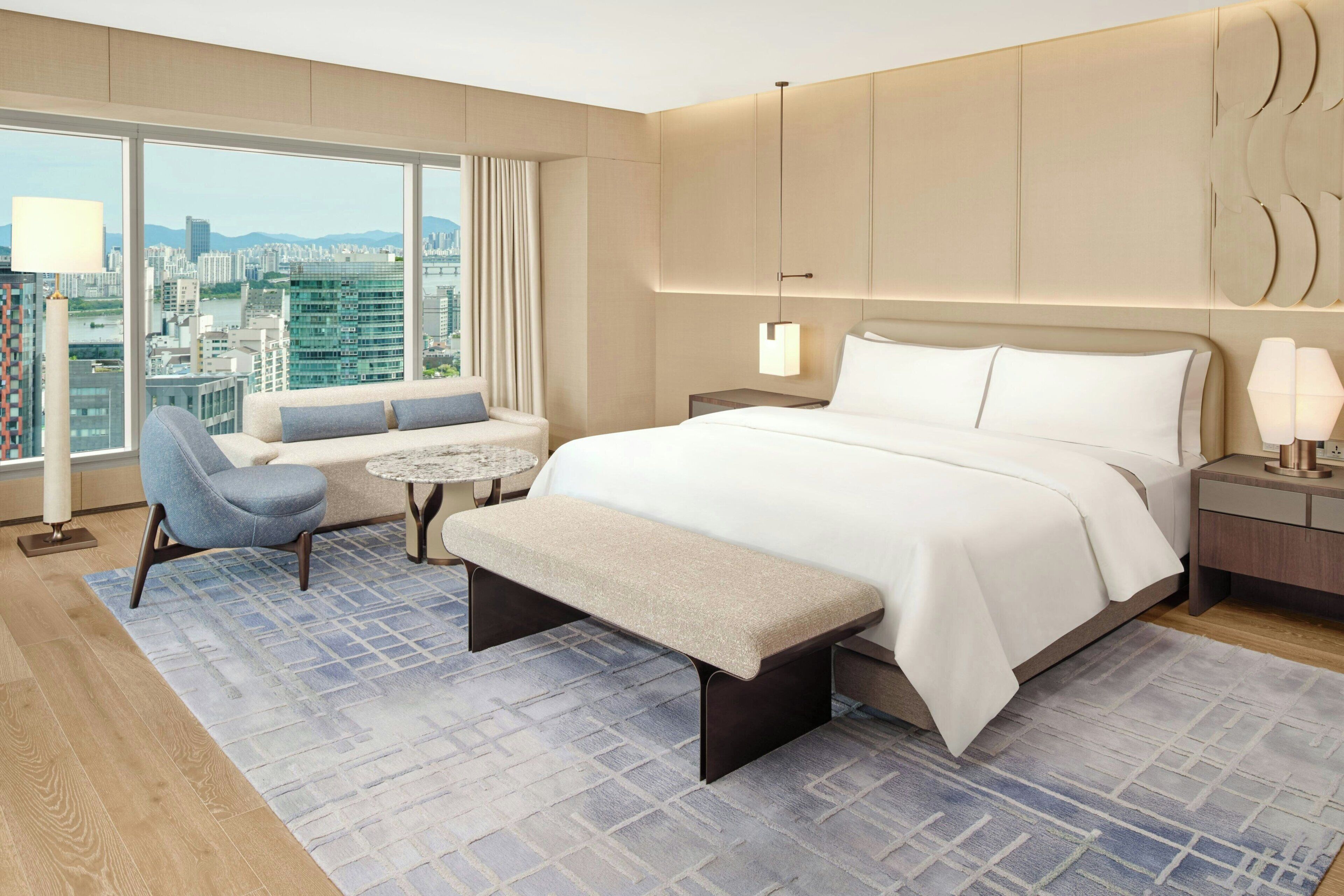 Una habitación de hotel moderna con una cama grande, una zona de estar con un banco, una mesita y vistas a la ciudad a través de amplios ventanales.