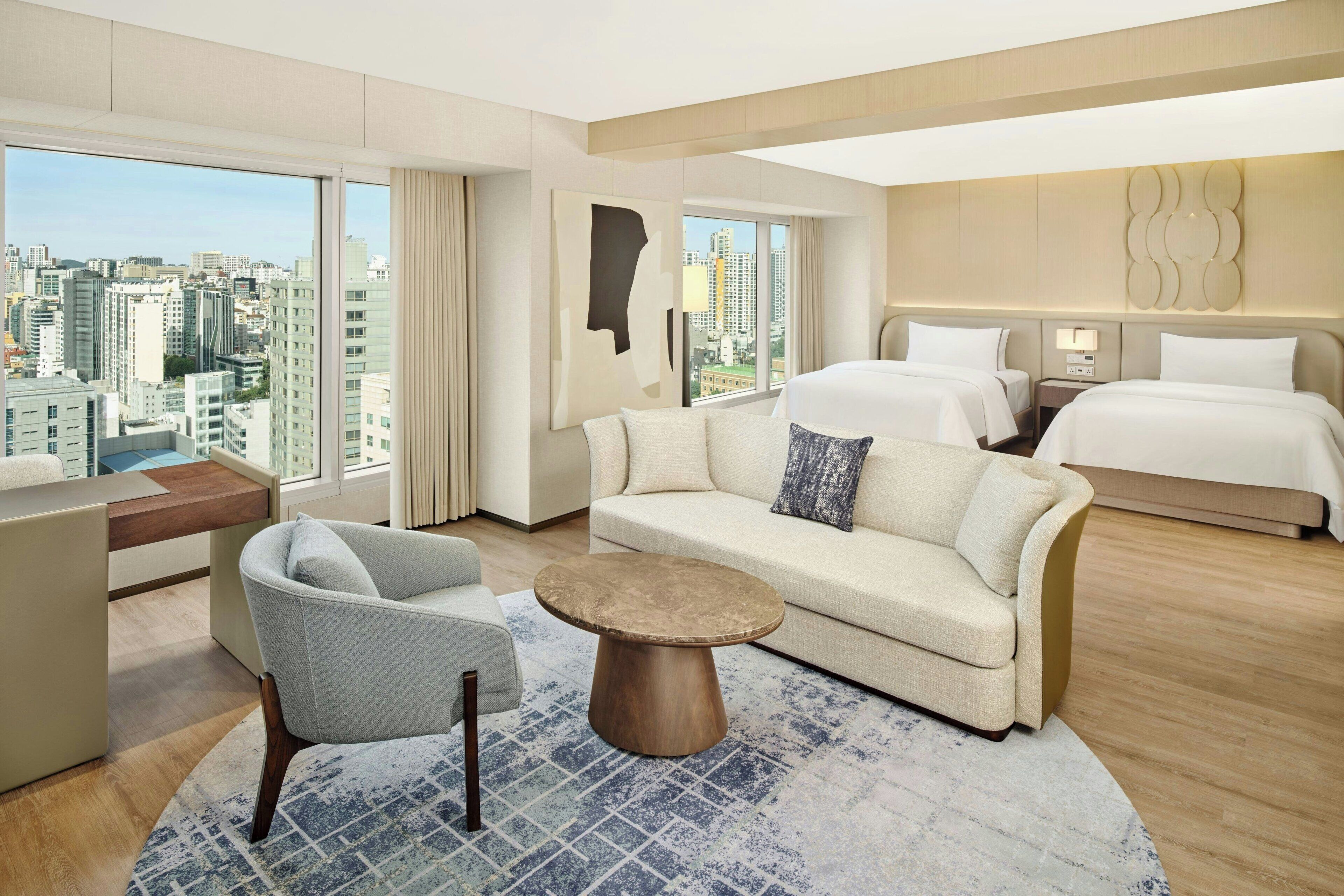 A modern hotel room with a city view, a sofa, a round coffee table, and a bed.
