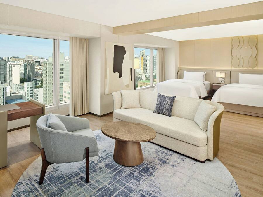 A modern hotel room with a city view, a sofa, a round coffee table, and a bed.