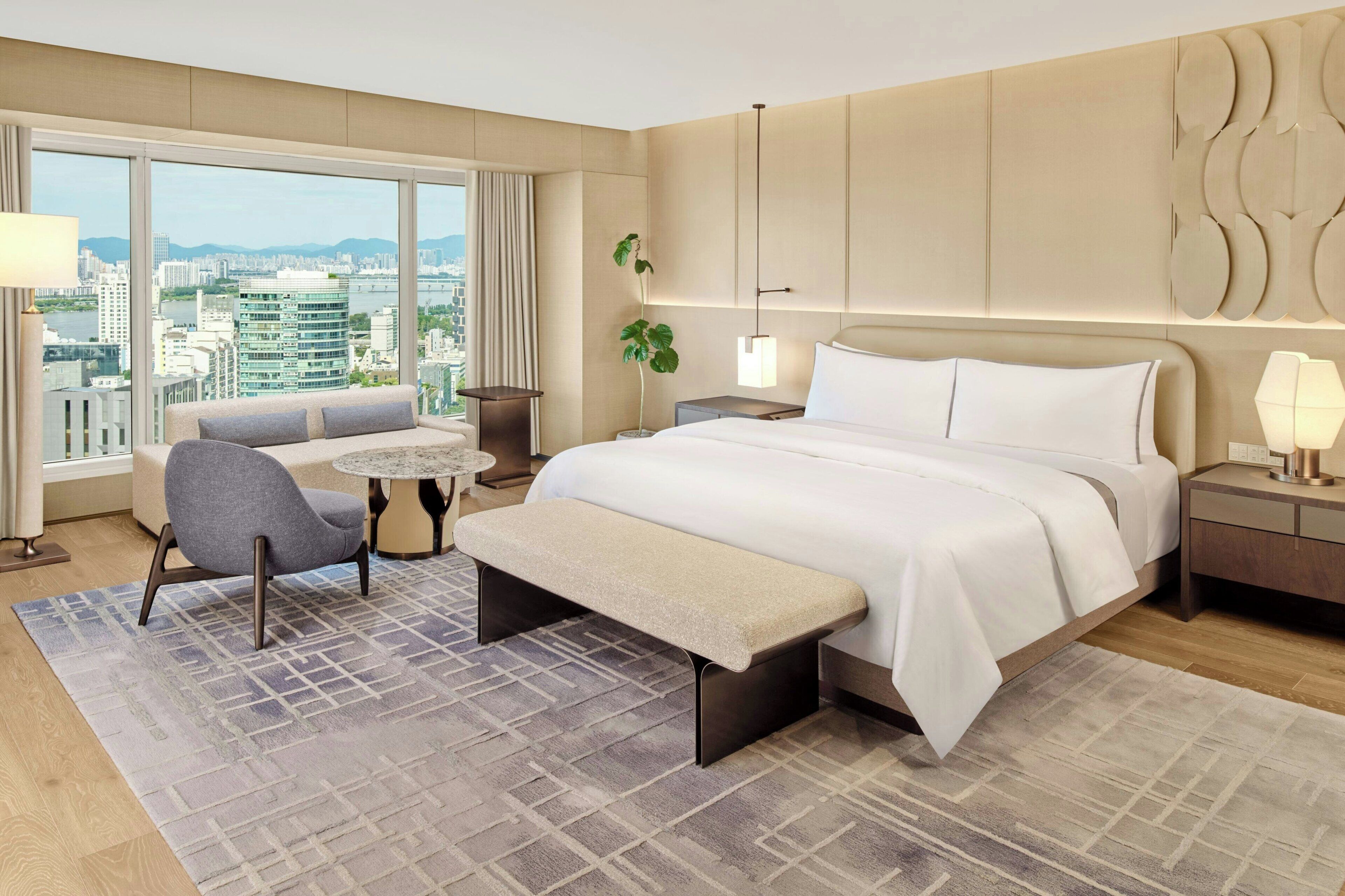 A modern hotel room with a large bed, a bench, a sofa, a small round table, and a view of the city.