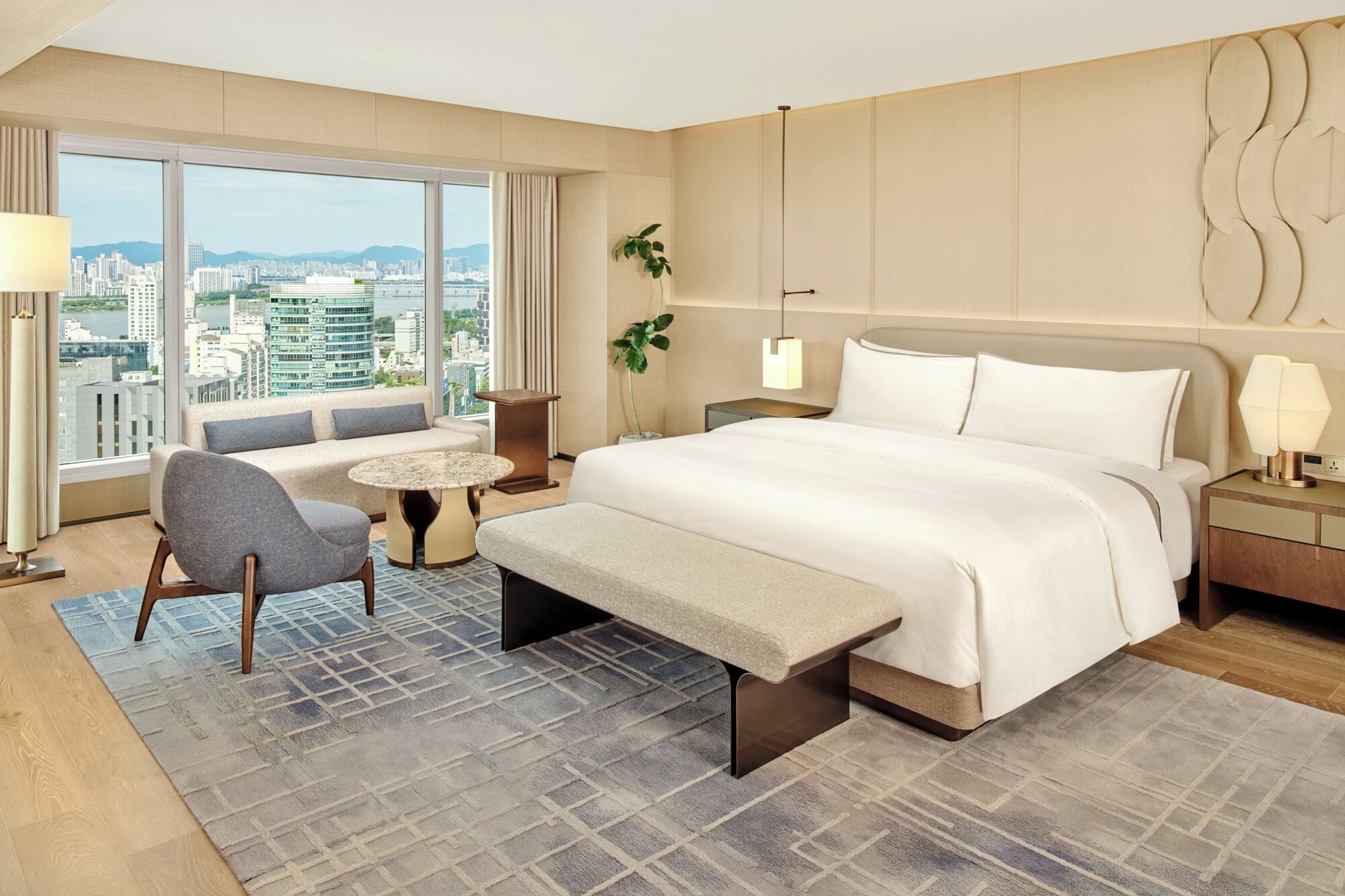 Habitación de hotel con una cama grande, un sofá, una mesita pequeña, una silla, una mesita de noche, una lámpara, una planta en maceta y vistas a la ciudad.