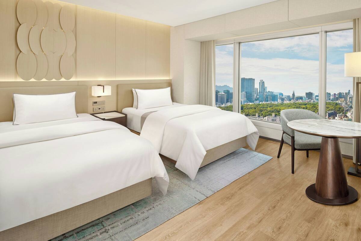 Une chambre d’hôtel avec deux lits, une table à manger, une chaise, une vue sur la ville et un décor mural moderne.