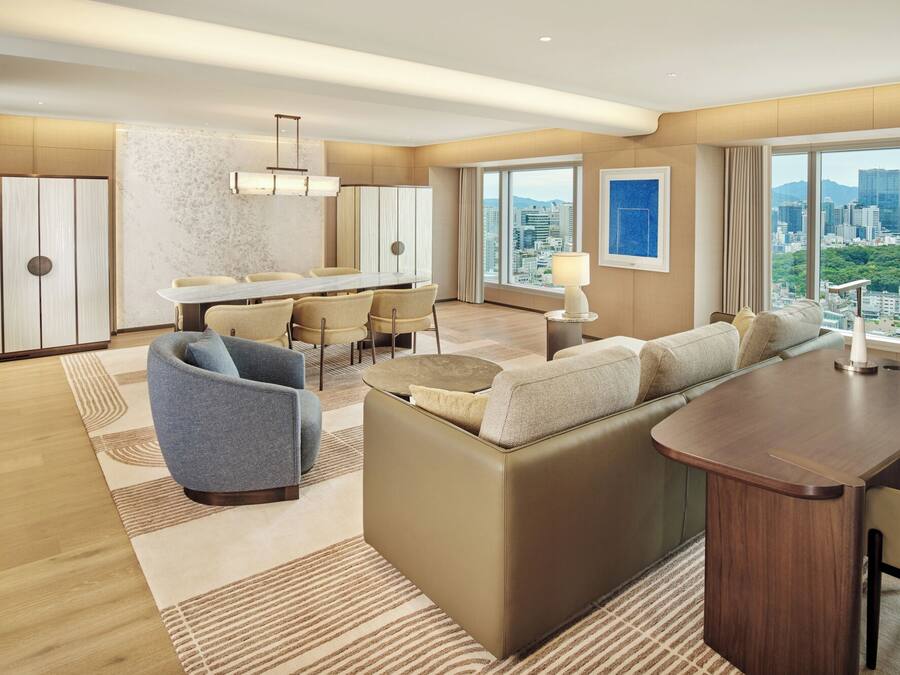 A modern hotel room with a dining area, a sofa set, a small table, and a view of the cityscape.